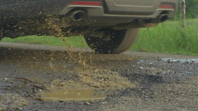 CLOSE UP: Car driving and splashing water from road pit puddle on paved road. Dangerous pothole with puddle in the middle of the asphalt road. Damage and bad road conditions in third world.