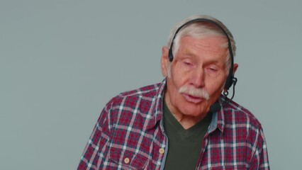 Senior old man wearing headset, freelance worker, call center or support service operator helpline, having talk with client or colleague, communication support. Elderly grandfather on gray background