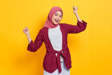 Excited beautiful Asian woman in casual shirt celebrating big luck and success with raised fists isolated over yellow background