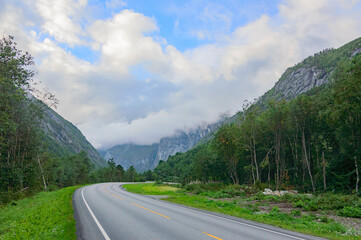 road to the mountains in Norway. Blue sky with clouds, grey asphalt, green trees and grass, mountain slopes far away.