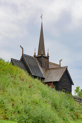 Fototapeta na wymiar Garmo Stave Church in Norway. Carved dragon heads on the roof, resembling those from the viking ships, dark ancient wooden walls, roof and bell tower, cloudy sky, green hill slope