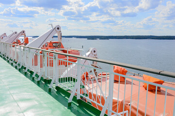 Upper deck of a cruise ship in a sunny day. Green floor, orange lifeboats, grey sea water, blue sky with clouds, green forest on a distant shore.