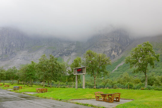 Norwegian summer. Weather screen at the Trollveggen rest area in Norway, showing +12 Celsius temperature. Green grass and trees, wooden tables and benhes, grey rocky slopes, low clouds in the sky.
