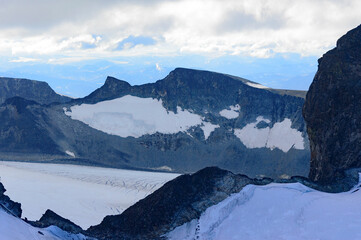 View of the mountain landscape in Jotunheimen national park in Norway from the Galdhopiggen mountain. Blue sky with clouds, grey and biege rocks, mountain slopes, white snow and ice of the glacier