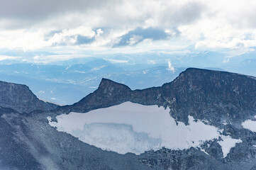 View of the mountain landscape in Jotunheimen national park in Norway from the Galdhopiggen mountain. Blue sky with clouds, grey and biege rocks, mountain slopes, white snow and ice of the glacier
