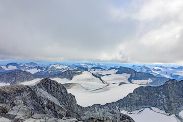 View of the mountain landscape in Jotunheimen national park in Norway from the Galdhopiggen mountain. Grey cloudy sky, grey and brown mountain slopes, white snow and ice of the glaciers