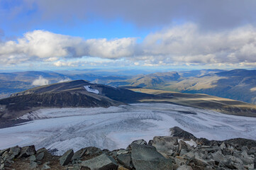 View of the mountain landscape in Jotunheimen national park in Norway from the Galdhopiggen mountain. Blue sky with clouds, grey and biege mountain slopes, white snow and ice of the glacier