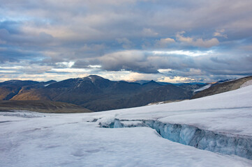 Evening on the glacier on the trail from Galdhopiggen summit in Norway. White, blue and grey cracked ice, dark mountains, sunset sky with clouds