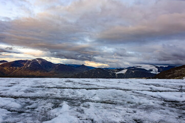 Evening on the glacier on the trail from Galdhopiggen summit in Norway. White and grey ice, dark mountains, sunset sky with clouds