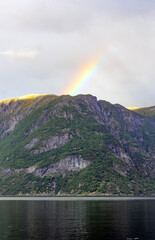 View of Geirangerfjord in Norway. Blue and grey sea water, blue sky with clouds, green and mounain slopes with sunshine and sadows on the forest, rainbow above the summit