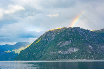 View of Geirangerfjord in Norway. Blue and grey sea water, blue sky with clouds, green and mounain slopes with sunshine and sadows on the forest, rainbow above the summit
