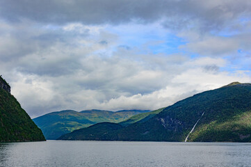 View of Geirangerfjord in Norway. Blue and grey sea water, blue sky with clouds, green and mounain slopes with sunshine and sadows on the forest, waterfall far away