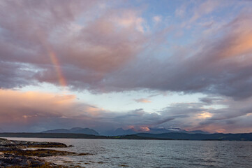 Norwegian landscape: blue evening sky with clouds  above the fjord, rainbow, calm water and dark mountains far away