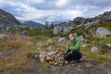 Young woman hiker with a harvest of boletus mushrooms in the northern Norwegian landscape. Grey mossy stones, blue mountains, cloudy sky