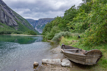 Old wooden boat on the river bank in Norway. Green grass and trees, clear water, mountains and cloudy sky on the background