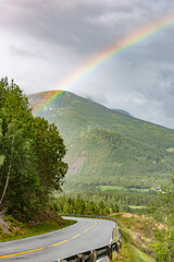 Norwegian scenery: rainbow in dramatic cloudy sky, asphalt road, green grass and trees and forest on mountain slopes in Eikesdal valley in Norway