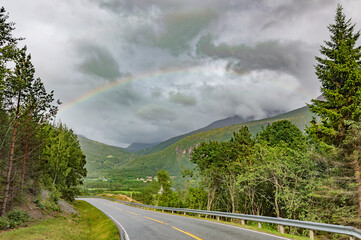 Norwegian scenery: rainbow in dramatic cloudy sky, asphalt road, green grass and trees and forest on mountain slopes in Eikesdal valley in Norway