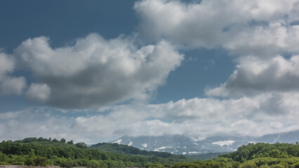 Fototapeta premium The top of the volcano is hidden in white cumulus clouds against a blue sky. Snow is visible on the slopes. Lush green vegetation in the valley. Kamchatka. Stratovolcano Vilyuchinsky