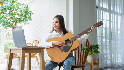 beautiful woman playing guitar happily.Cute Asian woman with attractive face spending free time playing music as  hobby.Thai female students studying songwriting online.Reading music notes on  laptop. - obrazy, fototapety, plakaty