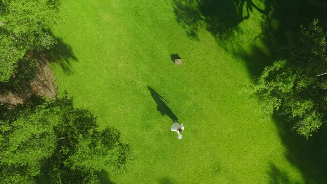 4K aerial footage energetic woman in white dress looking up in drone camera and taking off white hat on summer day. Happy young cheerful girl spinning around outdoors on green lawn Video series 1 of 6