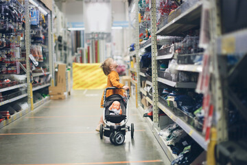 Cute baby girl in a mustard dress with doll in puppet stroller in a store, a child is playing in a hardware store