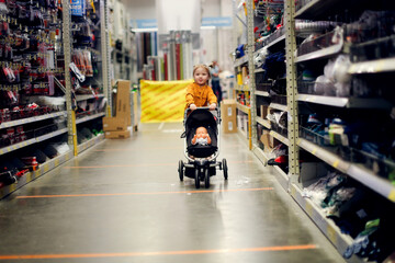 Cute baby girl in a mustard dress with doll in puppet stroller in a store, a child is playing in a hardware store