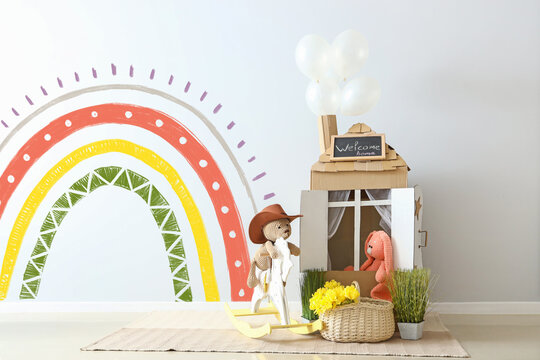 Toys, cardboard house and basket with flowers near light wall with painted rainbow