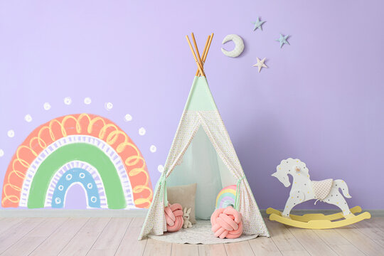 Children's wigwam, toys and rocking horse near wall with painted rainbow in room