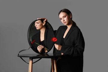 Fashionable woman standing near modern table with mirror and holding anthurium flower on grey background