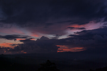 Colorful dark sunset landscape with bright orange, pink, yellow, purple, violet clouds on blue sky and silhouette dark hills in haze in Bali island, Munduk village.