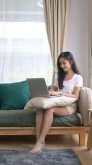 Fototapeta premium Full length portrait of happy young woman relaxing on sofa and browsing internet on laptop.