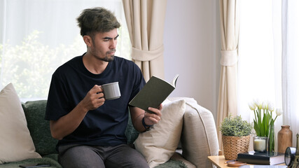 Peaceful millennial man drinking coffee and reading book on couch at home.