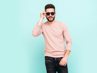 Portrait of handsome confident  model. Sexy stylish man dressed in pink sweater and jeans. Fashion hipster male posing near blue wall in studio. In sunglasses. Isolated