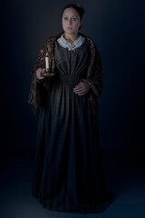Naklejka premium A Victorian working class woman standing alone holding a candle against a grey studio backdrop