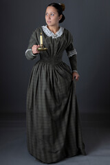 A Victorian working class woman standing alone holding a candle against a grey studio backdrop