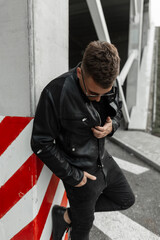 Fashion urban man model with hairstyle in black stylish leather jacket with sunglasses stands on the street