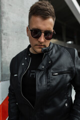 Fashion handsome male model with hairstyle and sunglasses in stylish leather black jacket on the street