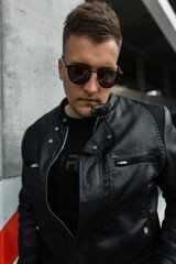 Obraz premium Fashion handsome male model with hairstyle and sunglasses in stylish leather black jacket on the street