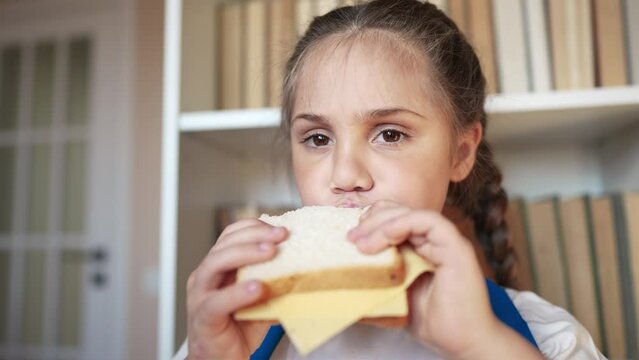 schoolgirl eats a sandwich at school during recess with backpack and bookcase. child lunch education concept. child in classroom having lunch snack lifestyle with bread and cheese sandwich