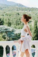 Portrait of young beautiful woman in a white shirt admires the green nature on the balcony sunny day