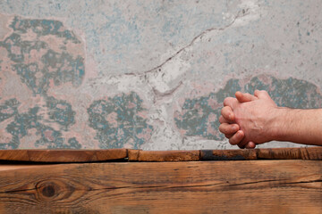 Intertwined male hands lying on the top of an old wooden table. In the background is a wall with faded color and deteriorated, cracked plaster.