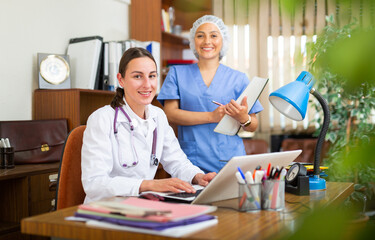 Confident woman doctor with assistant working in medical office using laptop computer, consulting patient online, telemedicine concept