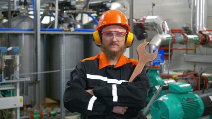 Technological mechanic in an orange helmet, headphones and goggles, holds a large wrench in his hands. Bearded male operator at the workplace in an industrial workshop.
