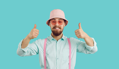 Cheerful funny charismatic young man showing thumbs up isolated on turquoise background. Bearded hipster in shirt, suspenders, panama and sunglasses looking at camera with funny expression on his face