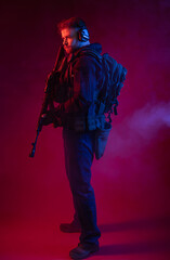Obraz premium soldier in full gear with weapons. a man in headphones, body armor, with a backpack and a belt. red background. colored, blue-red light