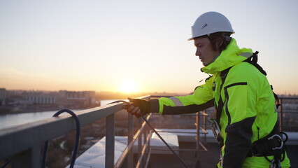 Industrial climber removes safety equipment from roof fence at sunset. Man in green overall...