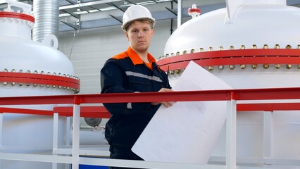 An engineer in a white helmet holds a drawing in his hands, checks the technological equipment and pipelines of gas and liquid, referring to the technological scheme. Factory engineer work