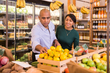 Obraz premium Hispanic man with his wife shopping together in grocery store, choosing ripe mandarines