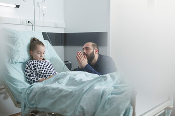 Young sad desperate father praying for sick little girl under treatment while in hospital bed....