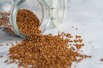 Close-up buckwheat grain poured from glass jar on gray table. Selective focus